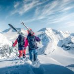 Freedride Kitz Two people with skis on their shoulders walking along a mountain in high snow.
